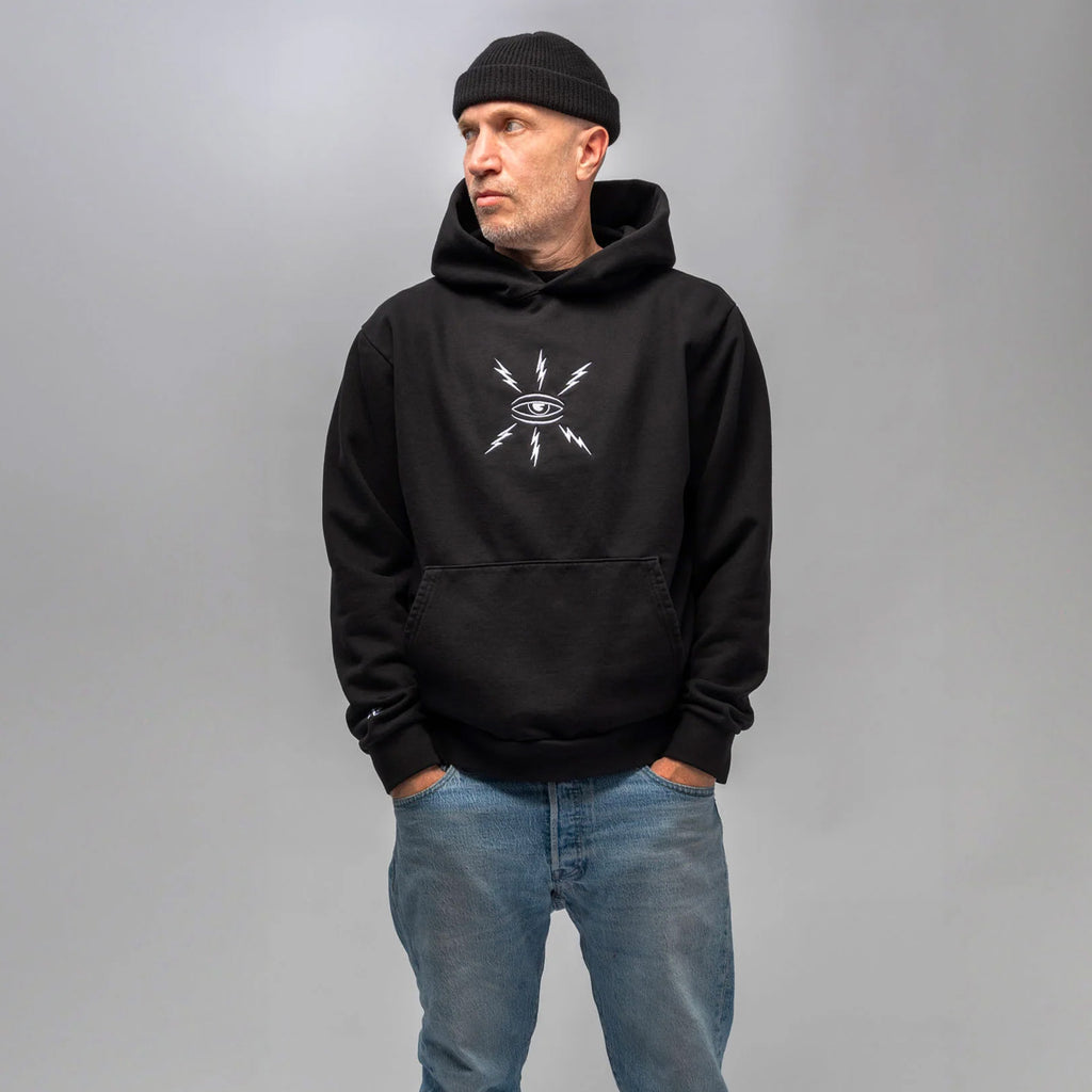 A man wearing the DICKIES WAR SAW X TEMPLETON COGNITIONS HOODIE BLACK stands against a gray background, paired with jeans and a black beanie, looking off to the side.