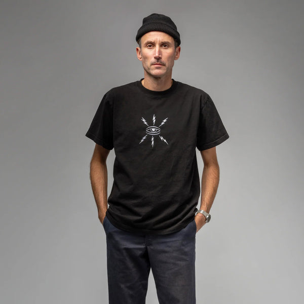 A man wearing a black beanie and the DICKIES WAR SAW X TEMPLETON COGNITIONS TEE, featuring an eye and lightning design, stands with his hands in his pockets against a gray background.