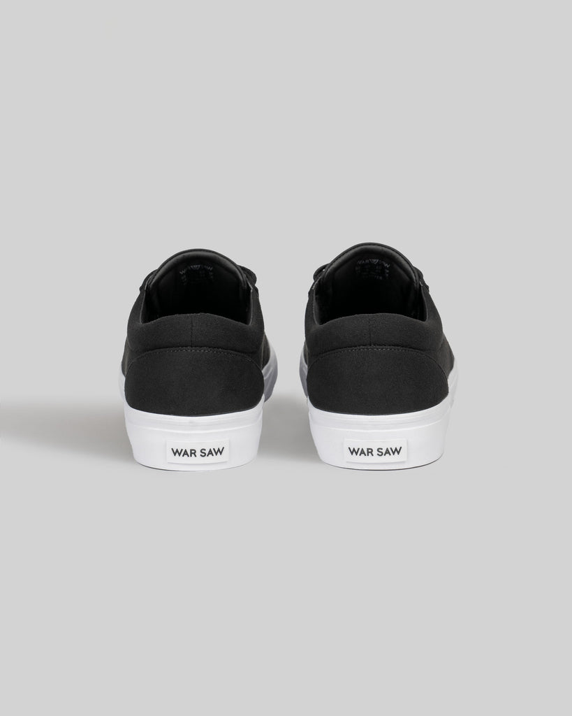 A pair of CHAPTER black and white synthetic sneakers by WARSAW LTD, skate-inspired with white soles, shown from the back and featuring "WAR SAW" on the heel tabs.