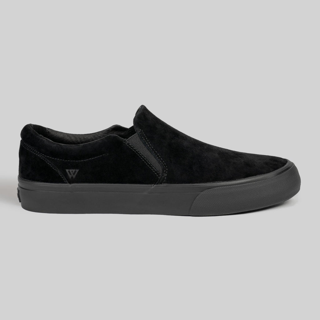 The STRANGER SLIP-ON | BLACK / BLACK by WARSAW LTD is a black genuine pig suede slip-on sneaker with a matching rubber sole and subtle stitching, shown against a plain gray background.