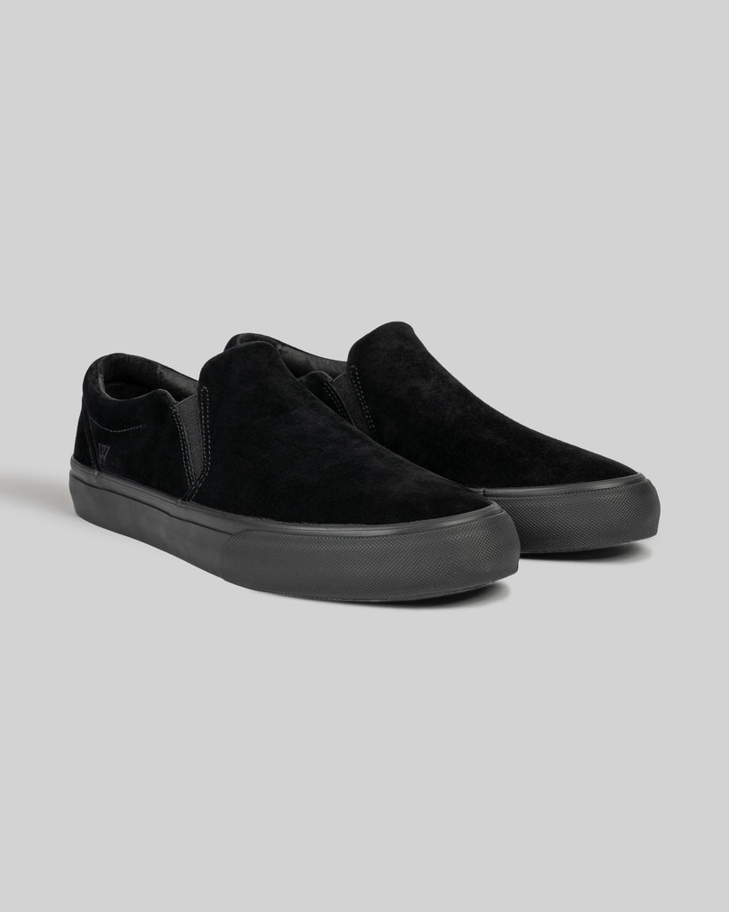 The WARSAW LTD STRANGER SLIP-ON | BLACK / BLACK features genuine pig suede uppers and rubber soles. These skate-inspired shoes are shown on a plain gray background.
