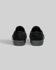 A pair of STRANGER SLIP-ON | BLACK / BLACK shoes by WARSAW LTD, made from genuine pig suede with "WARSAW" on the rubber soles, shown from the back against a plain gray background.