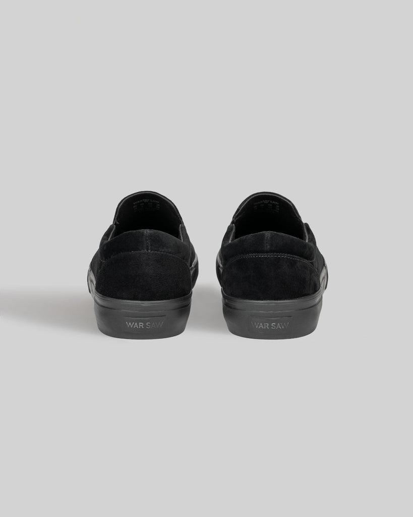 A pair of STRANGER SLIP-ON | BLACK / BLACK shoes by WARSAW LTD, made from genuine pig suede with "WARSAW" on the rubber soles, shown from the back against a plain gray background.