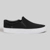 The WARSAW LTD STRANGER SLIP-ON | BLACK / WHITE SYNTHETIC is a black minimalistic slip-on sneaker with a white rubber sole, showcased against a plain light gray background.
