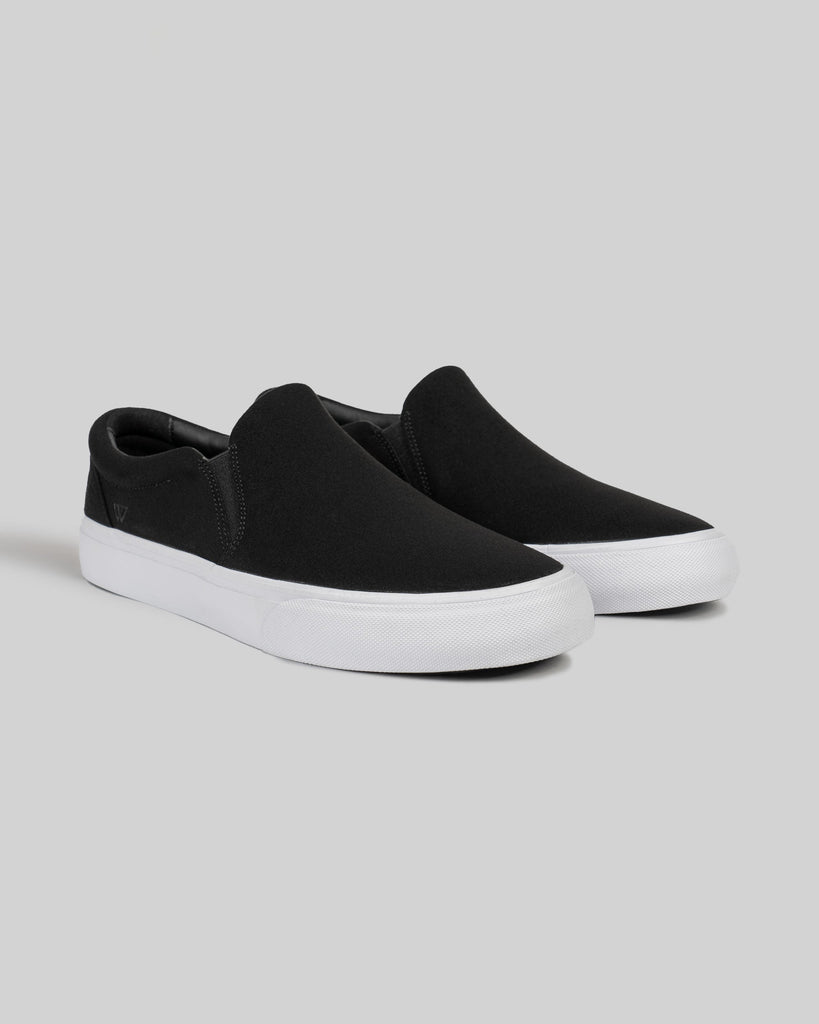 The WARSAW LTD STRANGER SLIP-ON in black/white synthetic features a minimalistic silhouette and white rubber soles, displayed on a plain grey background.