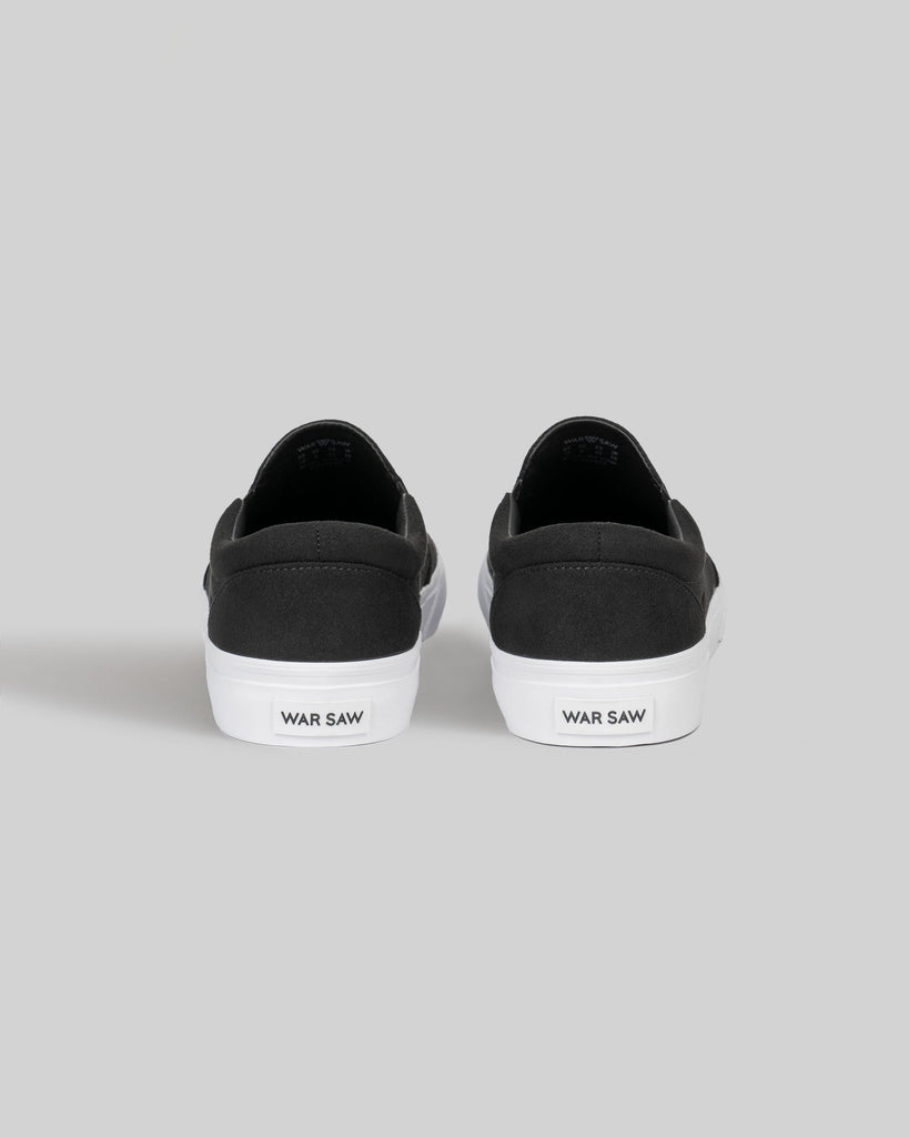 The STRANGER SLIP-ON | BLACK / WHITE SYNTHETIC by WARSAW LTD features black slip-on sneakers with white soles and "WARSAW" on each heel, showcasing a minimalistic skate-inspired design from the back.