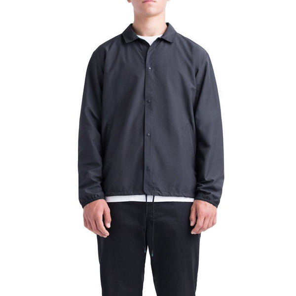 A person wearing the HERSCHEL SUPPLY CO VOYAGE COACH BLK jacket over a white shirt and black pants stands against a plain white background.