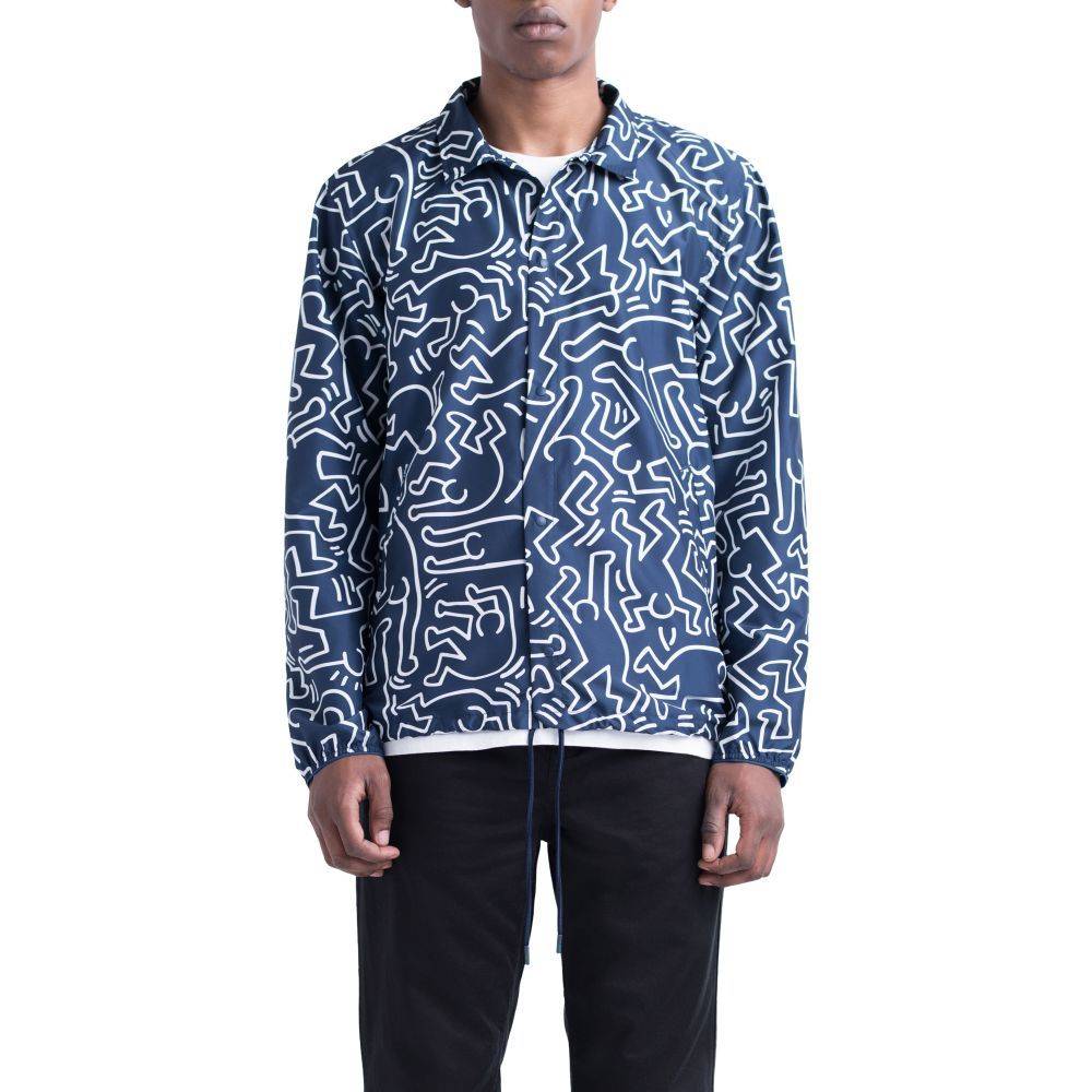 A person wearing the HERSCHEL X KEITH HARING VOYAGE COACH jacket, featuring a navy blue base with white abstract lines, paired with a white shirt and black pants, stands against a plain background.