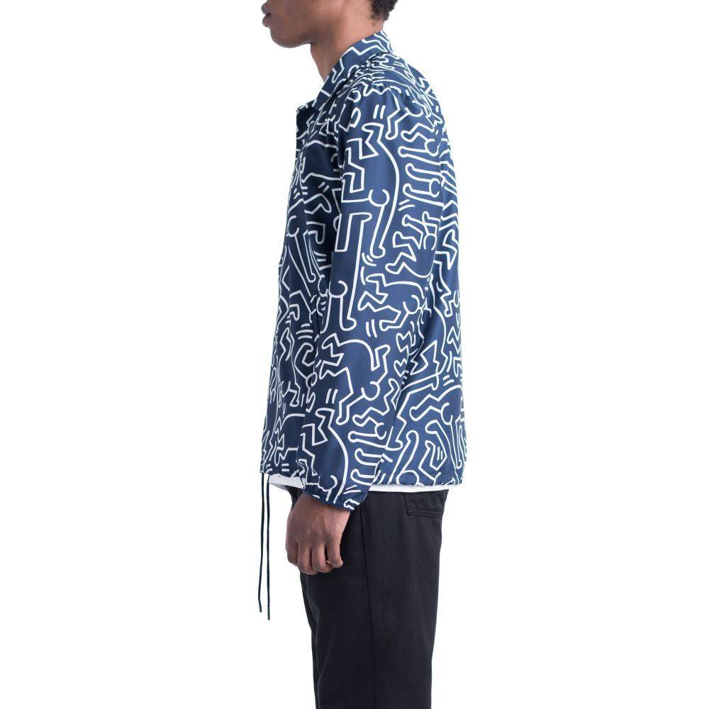 A person in side profile wears the HERSCHEL X KEITH HARING VOYAGE COACH jacket featuring a blue base with Keith Haring's white abstract line pattern, paired with black pants, against a plain background.