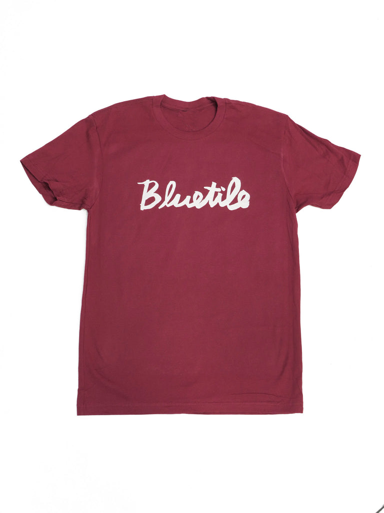 A Burgundy Bluetile Skateboards t-shirt with the word BLUETILE CURSIVE T-SHIRT MAROON / WHITE in cursive font.