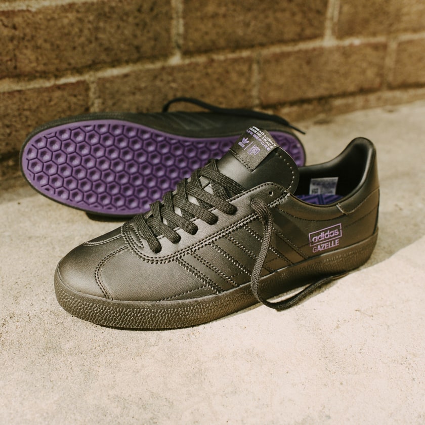 A pair of black ADIDAS PARADIGM GAZELLE BLACK / ACTIVE PURPLE sneakers on a brick wall.