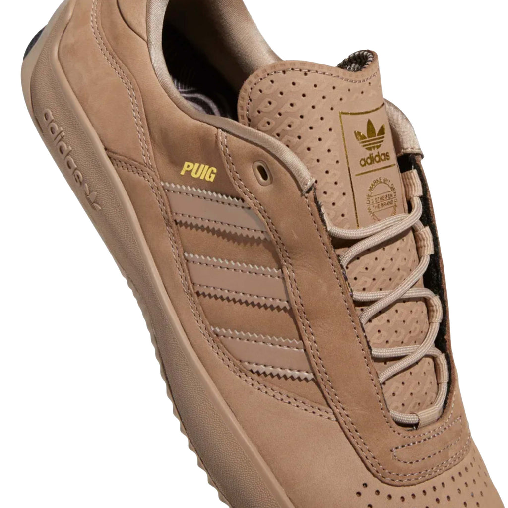 A pair of ADIDAS PUIG CHALKY BROWN / CORE BLACK shoes with gold accents in chalky brown.