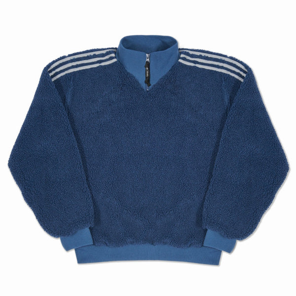 The ADIDAS X BLONDEY SHERPA CREW MINERAL BLUE pullover features a quarter-zip, ribbed cuffs and hem, and three white stripes on each shoulder—blending signature Blondey style with standout comfort and streetwear appeal.