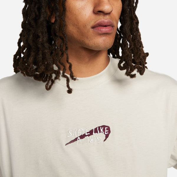 A person with long, twisted hair wears a light-colored nike NIKE SB x SKATE LIKE A GIRL MAX90 TEE featuring "SKATE LIKE A GIRL" embroidered above a maroon graphic.
