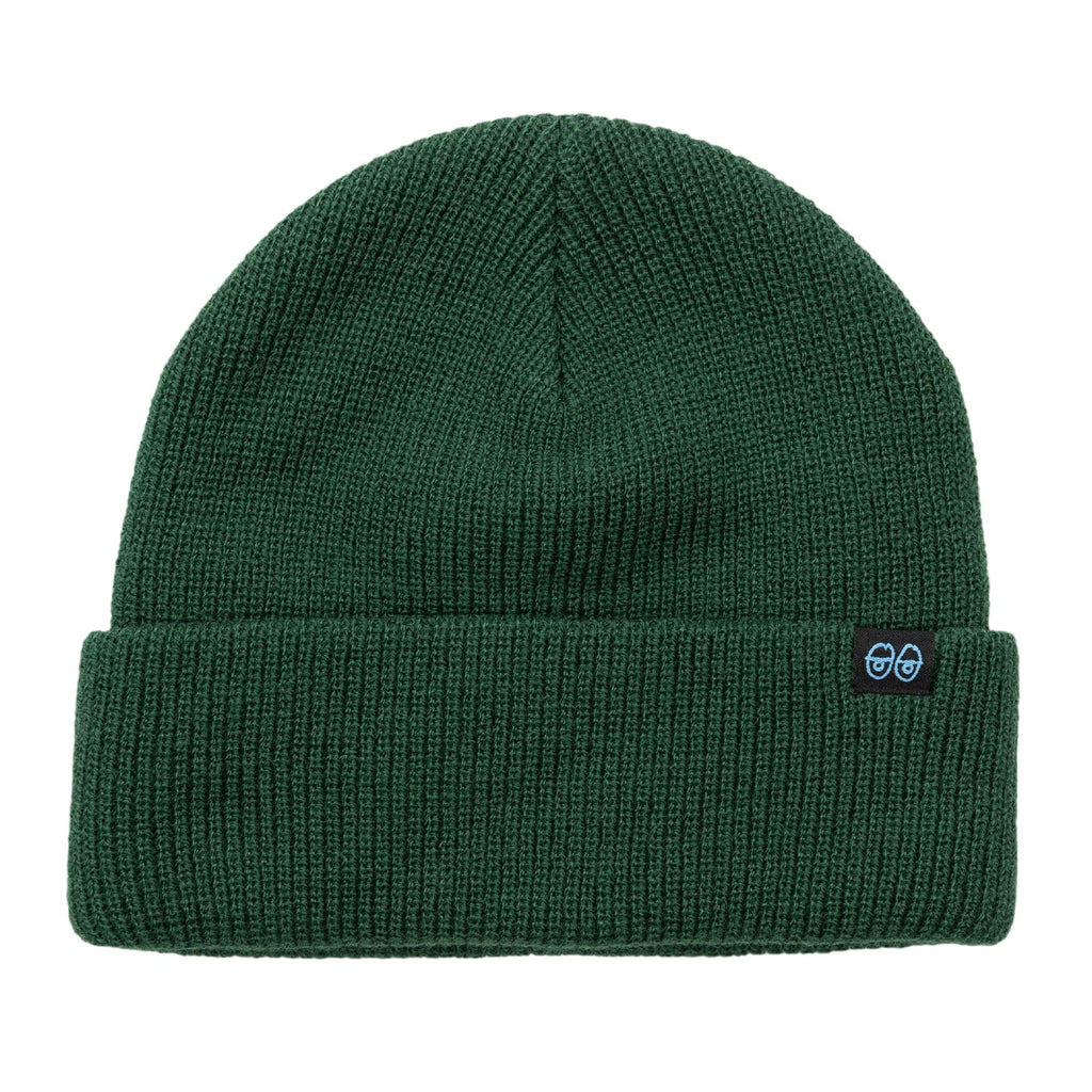 The KROOKED EYES CLIP CUFF BEANIE DARK GREEN/BLUE from DELUXE is a stylish and cozy dark green knit beanie featuring a folded brim and a small black patch with two blue illustrated faces.