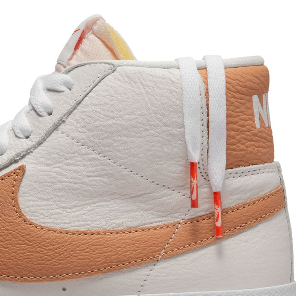 Close-up of a white and peach high-top NIKE SB BLAZER MID ORANGE LABEL WHITE / LT COGNAC sneaker with white laces and orange lace tips. The leather material showcases the nike logo on the side and back of the shoe.