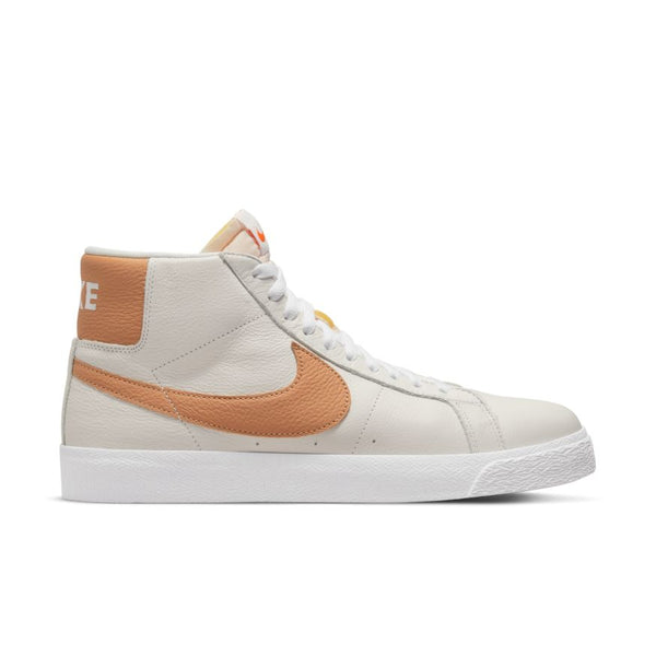 A white high-top sneaker in leather material with orange accents on the Nike swoosh and heel, featuring a white lace-up closure and a white rubber sole. Ideal for those who love skateboards, the NIKE SB BLAZER MID ORANGE LABEL WHITE / LT COGNAC combines style and function effortlessly.