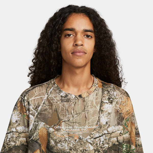 A person with long curly hair is wearing the NIKE SB x REALTREE® Print Skate Tee, featuring text and graphics on the front, standing against a plain background.