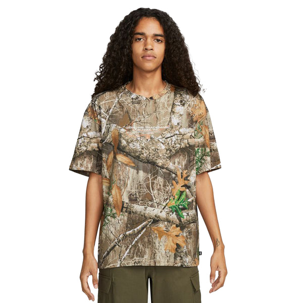 A person with long curly hair wearing a NIKE SB x REALTREE® PRINT SKATE TEE and green pants stands against a plain white background.