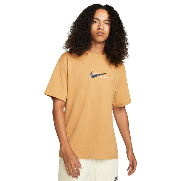 A person with long curly hair wears a tan NIKE SB EMBROIDERED SKATE TEE ELEMENTAL GOLD and light shorts, standing against a white background.