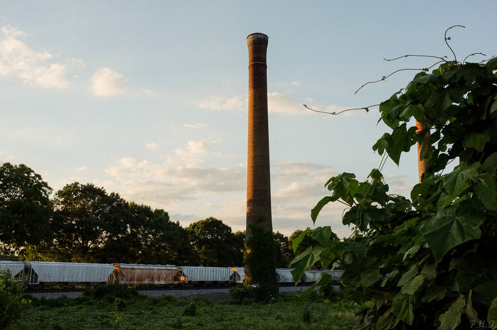 A Twin Train by Philip Taylor sits in a field next to a smokestack, captured in a mesmerizing photo print by Bluetile Skateboards.