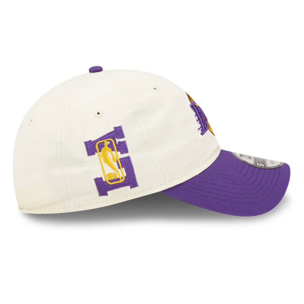The NEW ERA LOS ANGELES LAKERS NBA DRAFT CAP is a white baseball cap with a purple brim, featuring the Los Angeles Lakers' NBA Championship trophy graphic and the letter "L" on the side.