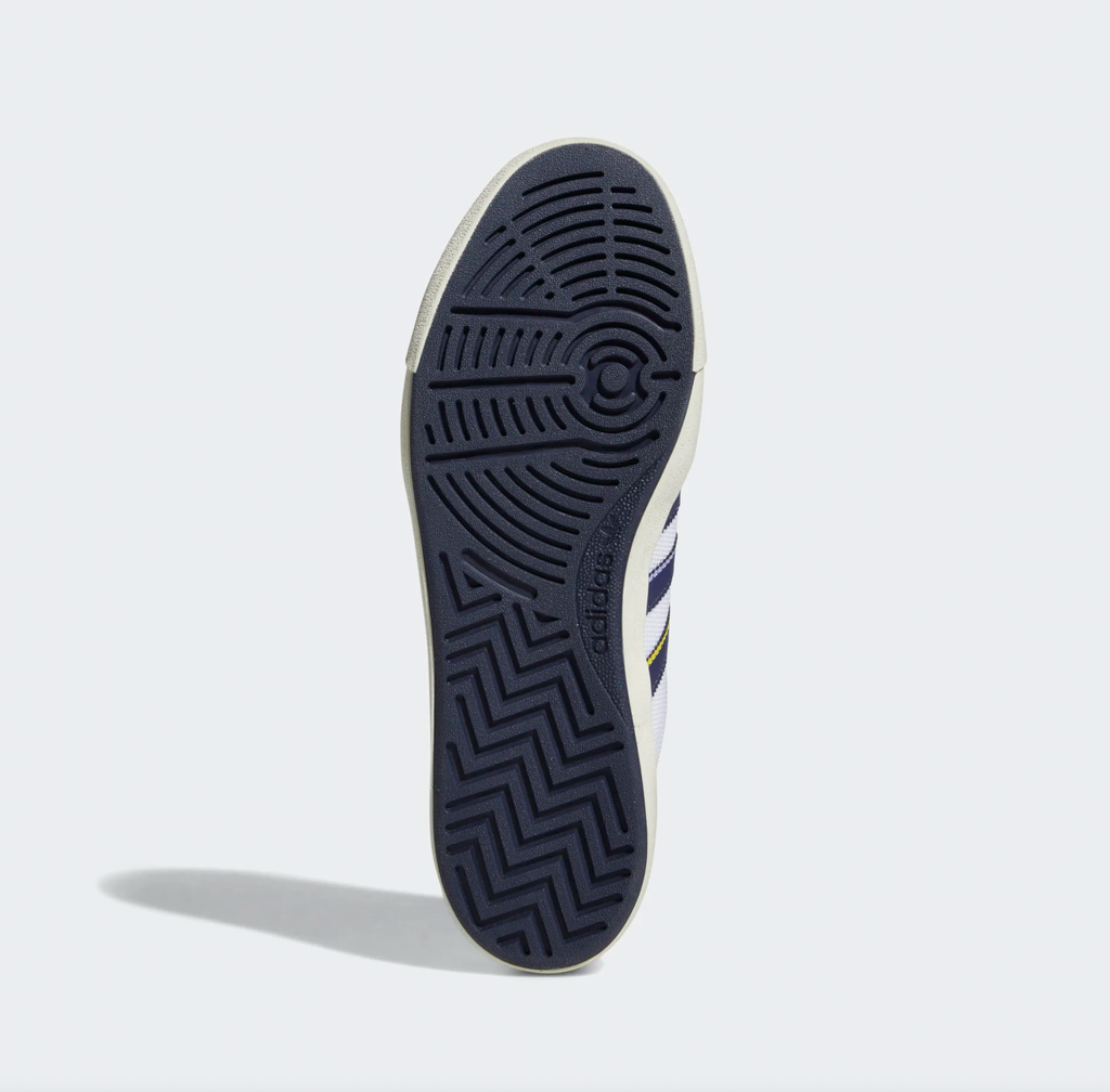 A close up of a person's ADIDAS Nora Grey One/ Shadow Navy / Gold shoe on a white background.