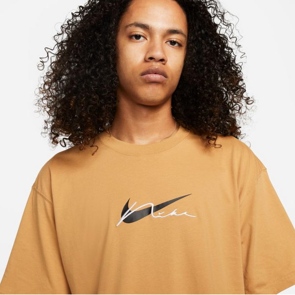 Person with curly hair wearing a Nike SB Embroidered Skate Tee Elemental Gold by Nike, featuring a stylized black swoosh and white "Nike SB" text.