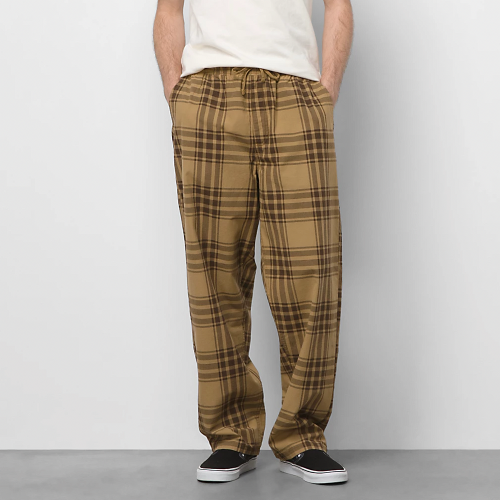 A man standing in front of a white wall wearing VANS RANGE PLAID BAGGY TAPERED ELASTIC WAIST PANT BROWN.