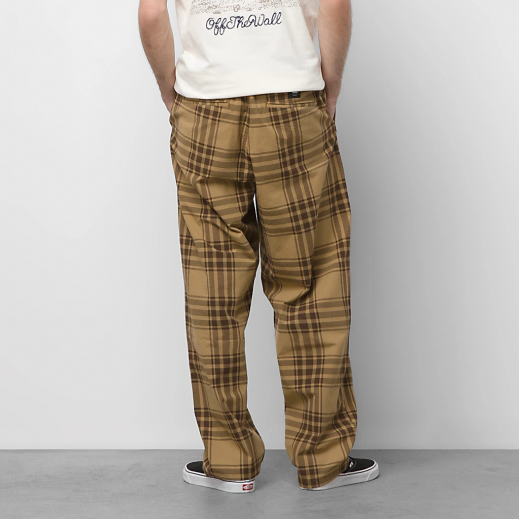 A man standing in front of a white wall wearing VANS RANGE PLAID BAGGY TAPERED ELASTIC WAIST PANT BROWN.