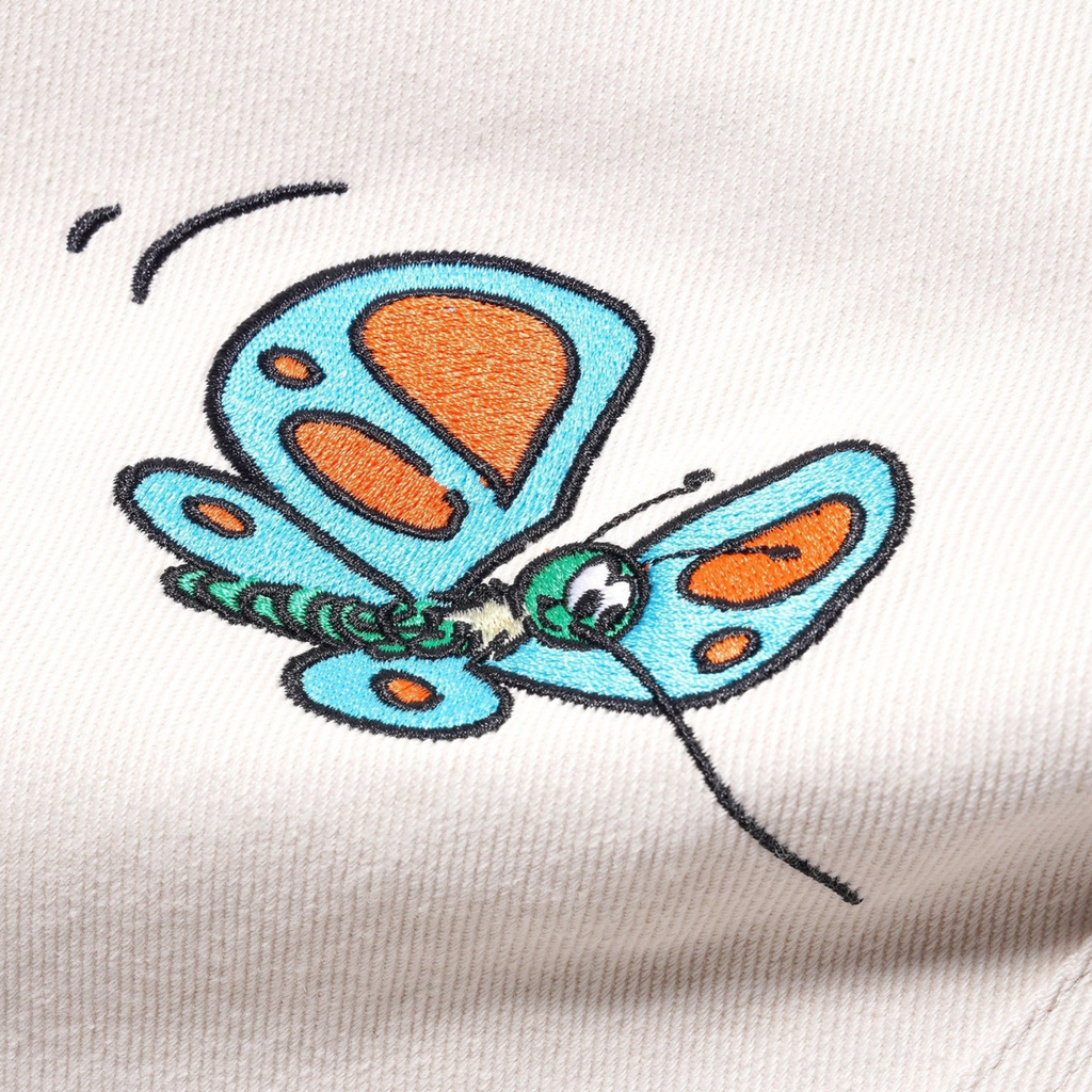 A close up of a blue butterfly on a Butter Goods X The Smurfs Forage Wide Leg Pant Natural from Butter Goods shirt.