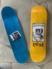 Two skateboards on a stone surface. The left skateboard is blue, featuring an image of a computer monitor and the words "test group." The right skateboard, designed with twin tail graphics, is yellow with a drawing of a burning monitor and the brand name "TEST FIREWALL TWIN TAIL" by TEST.