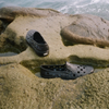 A pair of VANS TREK SLIP ON BLACK shoes sitting on top of a rock next to the ocean.