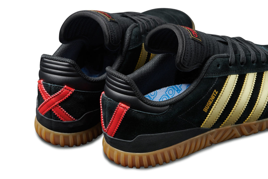 A pair of ADIDAS BUSENITZ INDOOR SUPER BLACK/GOLD/SCARLET sneakers in black and gold.