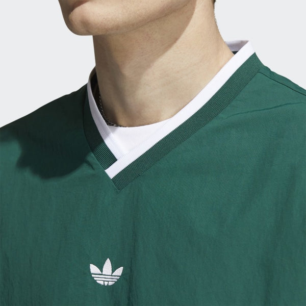 A person wears the ADIDAS WIND PULLOVER COLLEGIATE GREEN / WHITE, featuring a white logo and V-neck collar. The image spotlights the upper chest and neck, emphasizing its sporty, vintage golf windbreaker style.