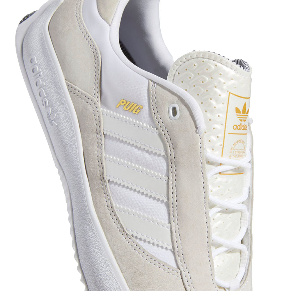 An Adidas Puig Cloud White/Core Black sneaker with a gold logo on the side.