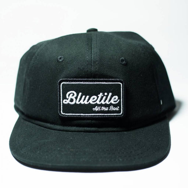 A black hat with a white patch on it, resembling the BLUETILE "ALL THE BEST" PATCH BLACK look from Bluetile Skateboards.