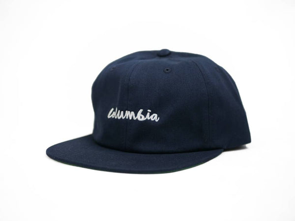 The CHOCOLATE CHUNK OF COLUMBIA HAT by CHOCOLATE features a navy flat brim and "columbia" embroidered in white lowercase letters on the front—combining classic style with comfort.