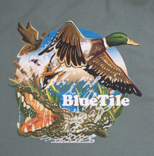 A Bluetile Skateboards BLUETILE WILDERNESS DUCKS T-SHIRT CYPRESS GREEN with an alligator and a duck flying in the air.