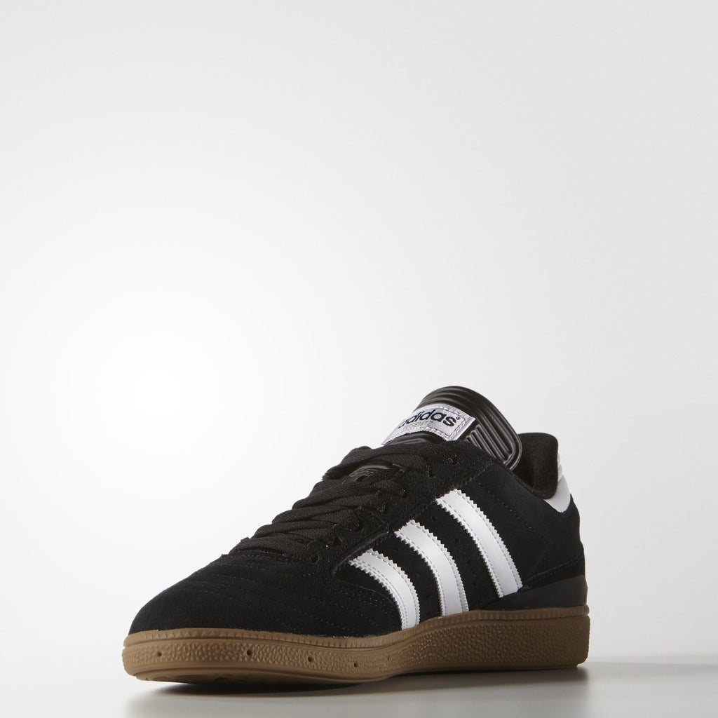A pair of ADIDAS BUSENITZ CORE BLACK / WHITE / GOLD sneakers on a white surface.