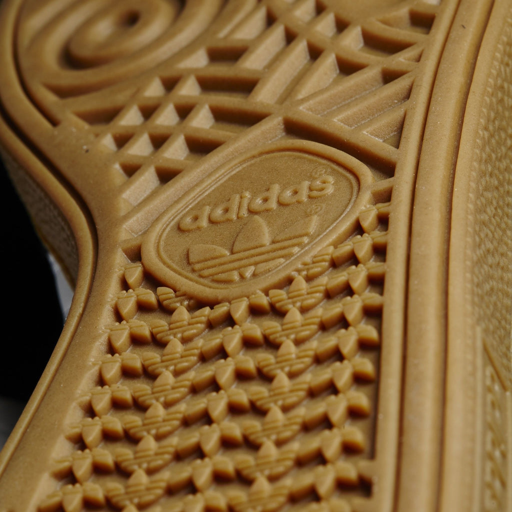 A close up view of the sole of an ADIDAS BUSENITZ CORE BLACK / WHITE / GOLD shoe.