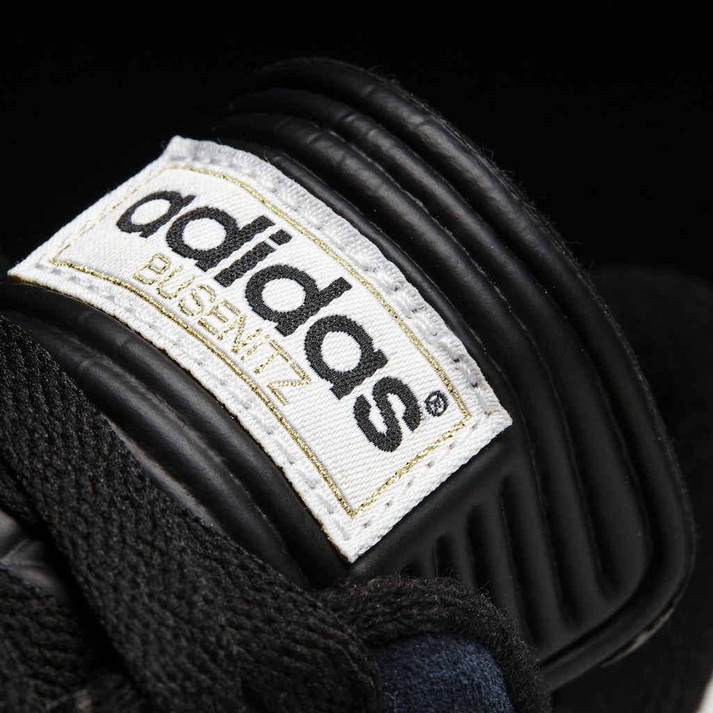 A close-up of an Adidas Busenitz Core Black / White / Gold shoe with a label on it.