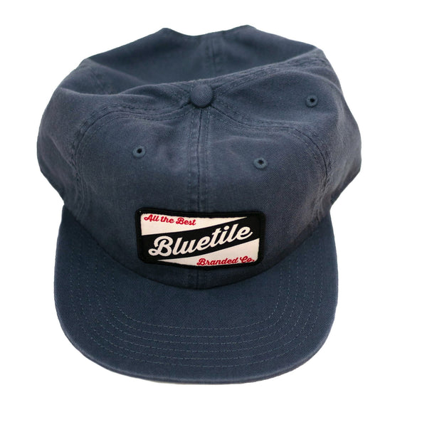 Description: A navy blue BLUETILE CRAFT PATCH HAT PETROL BLUE hat with the word bluetie on it, featuring a Bluetile Skateboards patch.