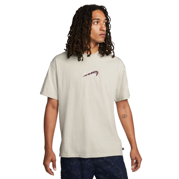 A person with long curly hair wears a light beige NIKE SB x SKATE LIKE A GIRL MAX90 TEE by nike, featuring a small Nike logo and dark patterned shorts, standing against a plain white background.