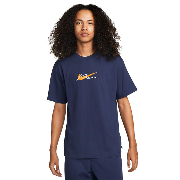 A person with long curly hair wears a midnight navy NIKE SB Embroidered Skate Tee by Nike, featuring an embroidered orange and white logo on the chest, standing against a white background.