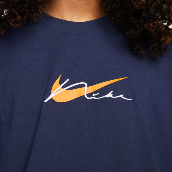 A person wears a NIKE SB Embroidered Skate Tee in midnight navy by nike, featuring an orange swoosh and "Nike" in white script overlapping the logo.