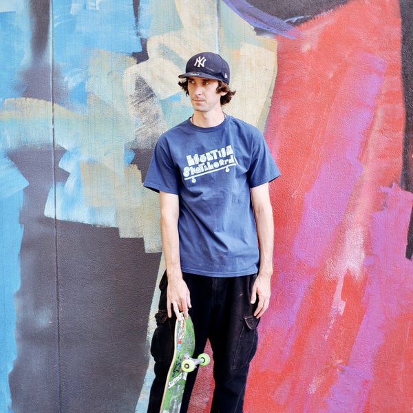 A man holding a skateboard in front of a colorful wall, wearing a Navy basic shapes tee.