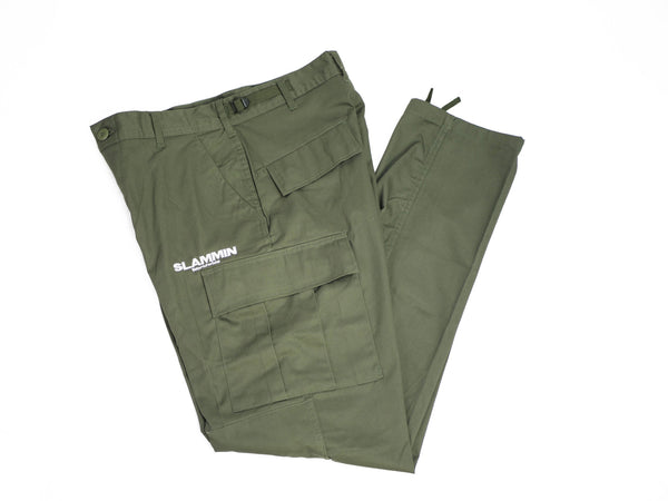 The SLAMMIN CARGO PANT OLIVE, neatly folded, features multiple pockets with the word "SLAMMIN" printed in white on one of the cargo pockets. Designed for durability with a reinforced seat and knees, these pants include adjustable waist tabs and multi-use utility pockets for added functionality.