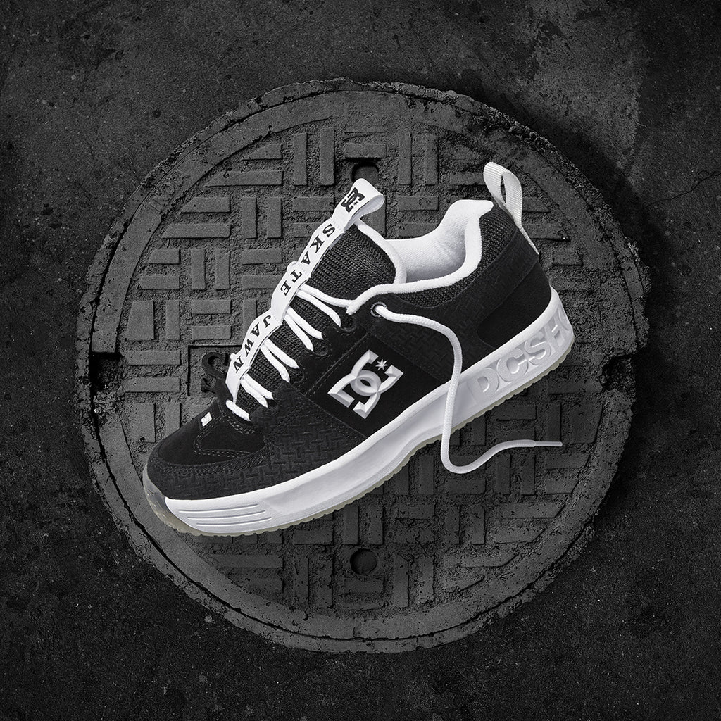 A black and white DC LYNX OG SKATE JAWN on top of a manhole, attracting the attention of skateboarders passing by.