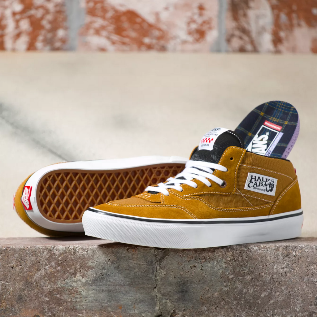 The VANS REYNOLDS SKATE HALF CAB '92 GOLDEN BROWN is sitting on top of a brick wall, showcasing the iconic skate shoe in its signature golden brown colorway.